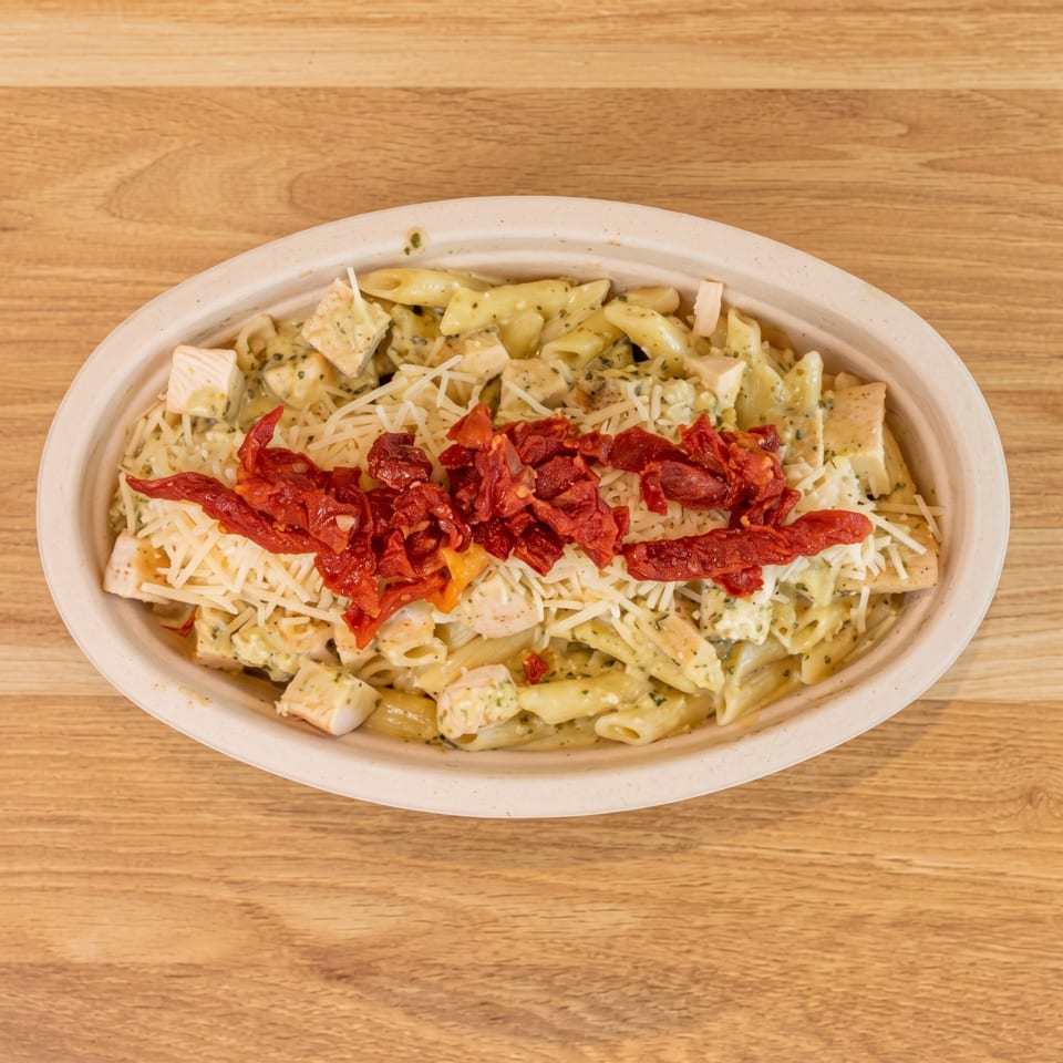 Penne pasta with chicken, pesto, shredded cheese, and sun-dried tomatoes in a disposable bowl.