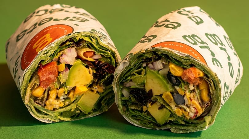 Two Greta wraps, filled with avocado, corn, tomatoes, and greens, on a green background.
