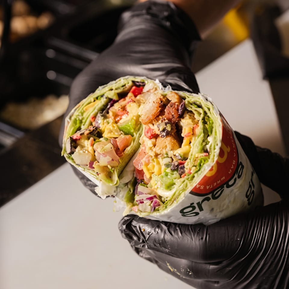 Gloved hands hold a Greca wrap, cut in half to reveal colorful fillings on a cutting board.