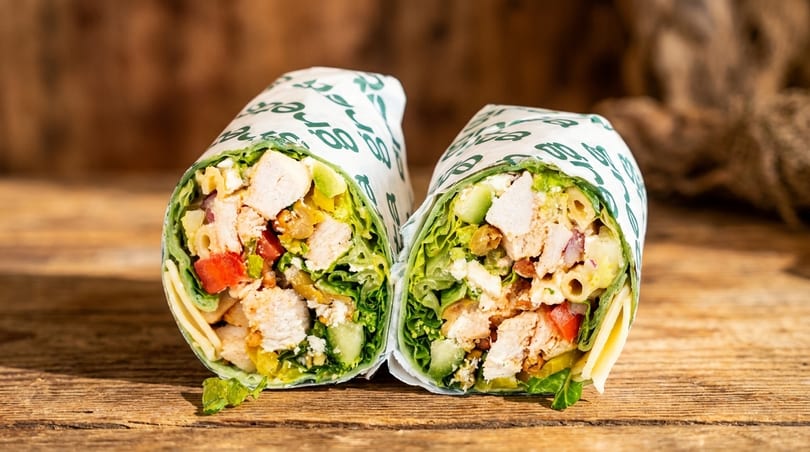 Two halves of a chicken wrap, filled with lettuce, tomatoes, and pasta, on a wooden surface.