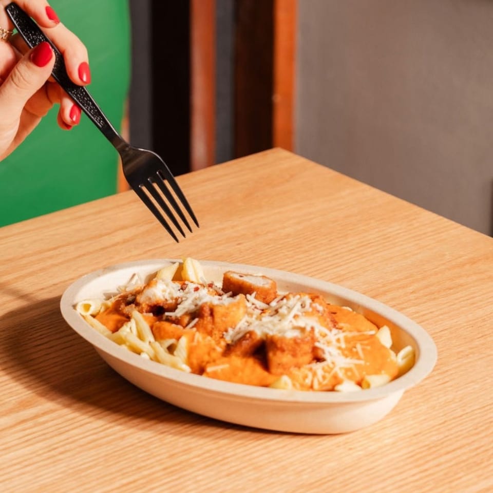 Hand with red nails holds a fork over penne pasta with creamy tomato sauce, chicken, and cheese.